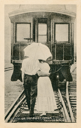 Vintage photograph titled "On the Right Track, Kenoza Lake, N. Y." showing a hugging couple under an umbrella while walking along the train tracks.