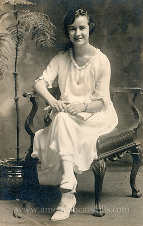 Otto Hillig undated portrait of a young woman, seated, smiling, white dress, hands crossed with book, plant in background.
